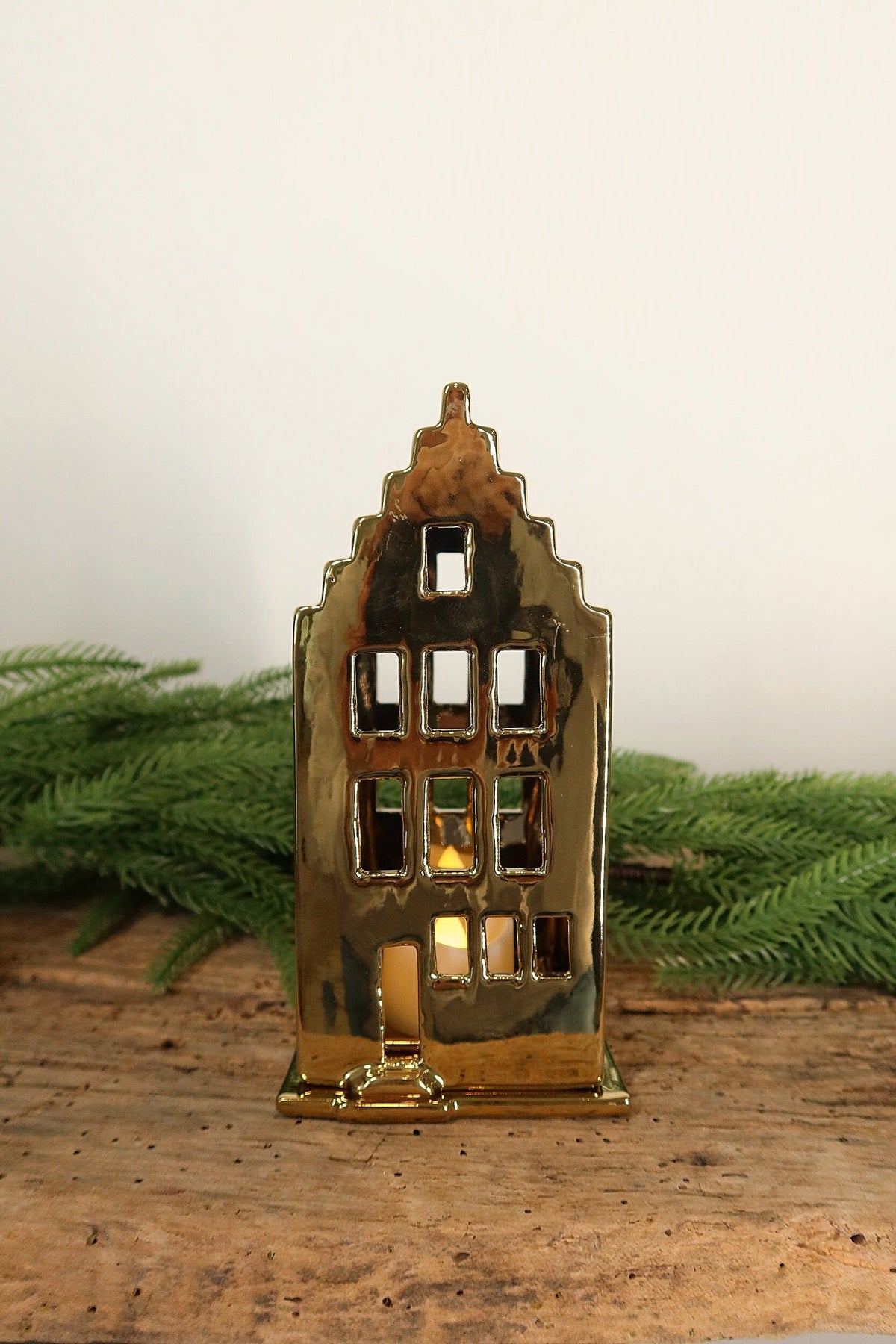 Gold ceramic canal house tealight holder glowing on a rustic wood table with greenery