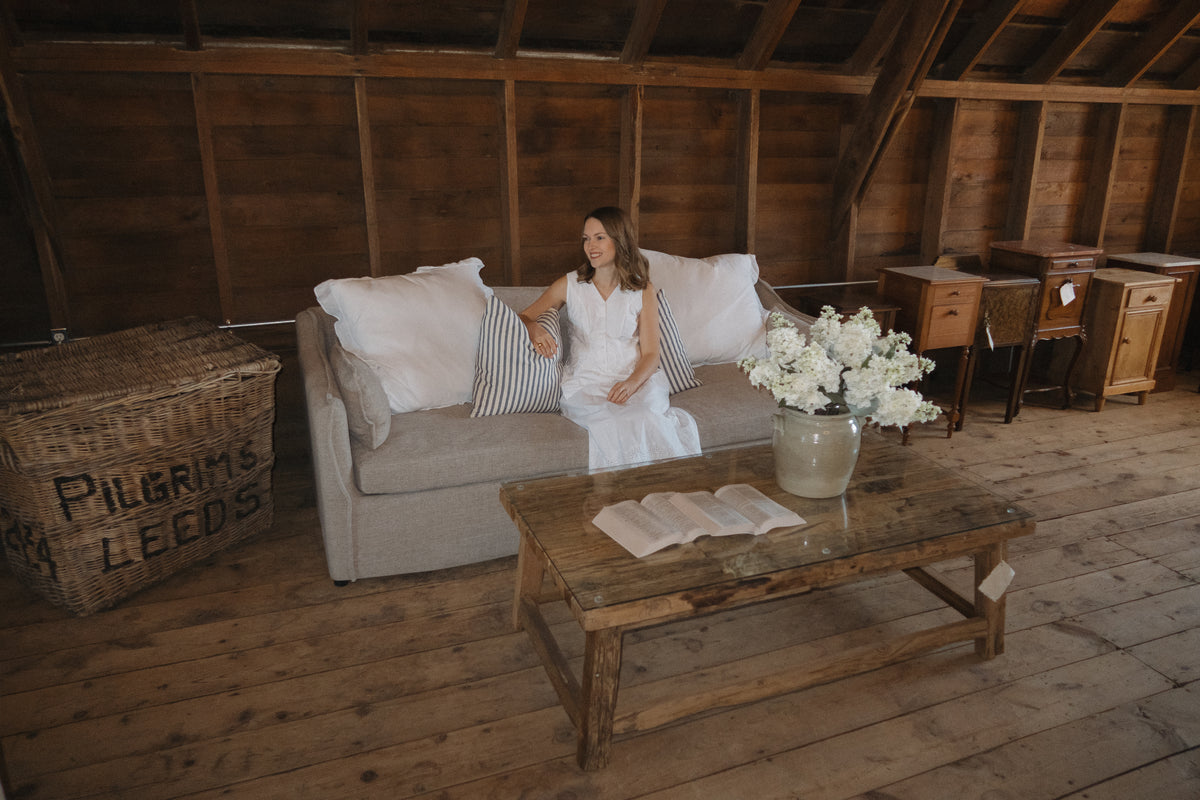 A woman relaxes on a linen sofa beside an antique wood coffee table and a vintage Pilgrim Leeds basket in the barn loft