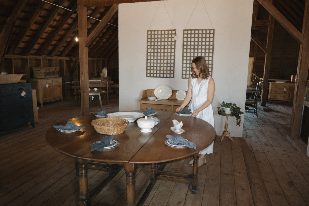 A woman sets a round antique table with white ironstone and blue linen napkins inside the Marvella Mercantile barn