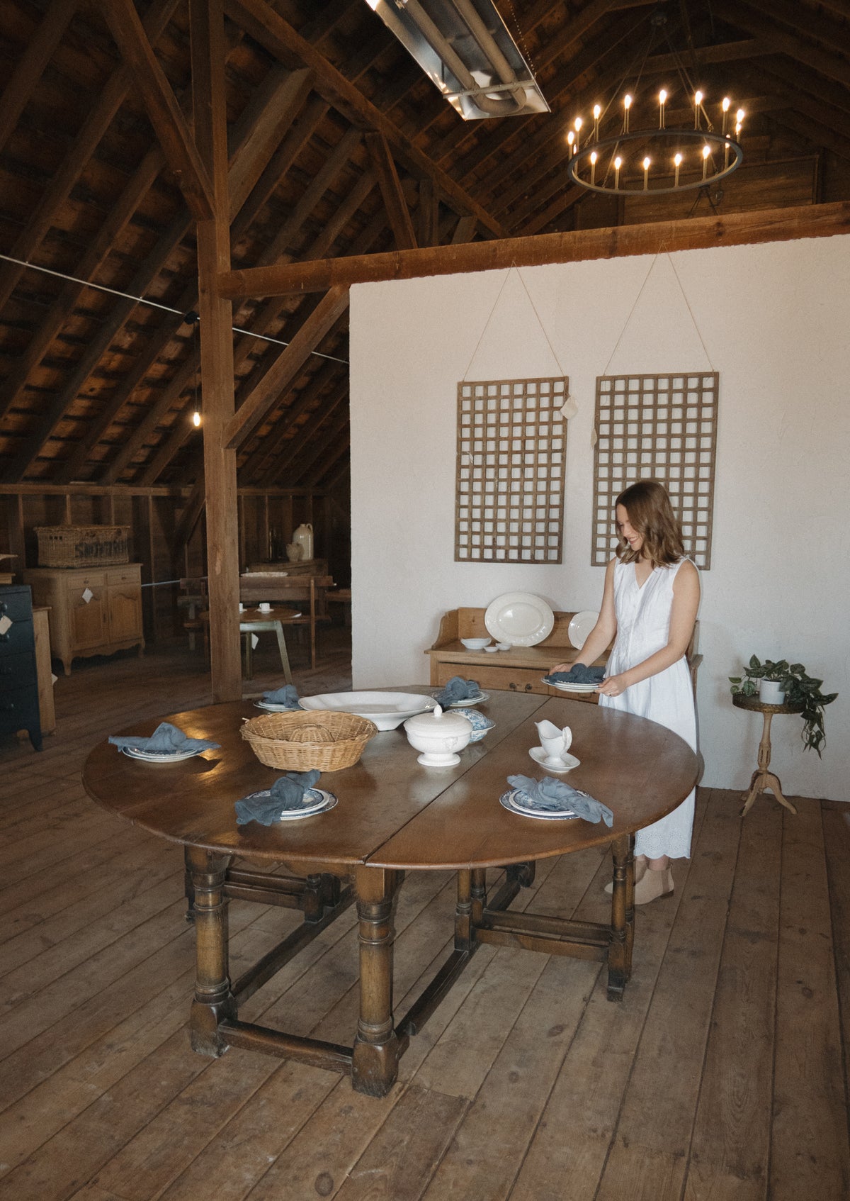 A woman sets a round wooden table with white ironstone and blue linen napkins beneath a candle chandelier in the timbered barn