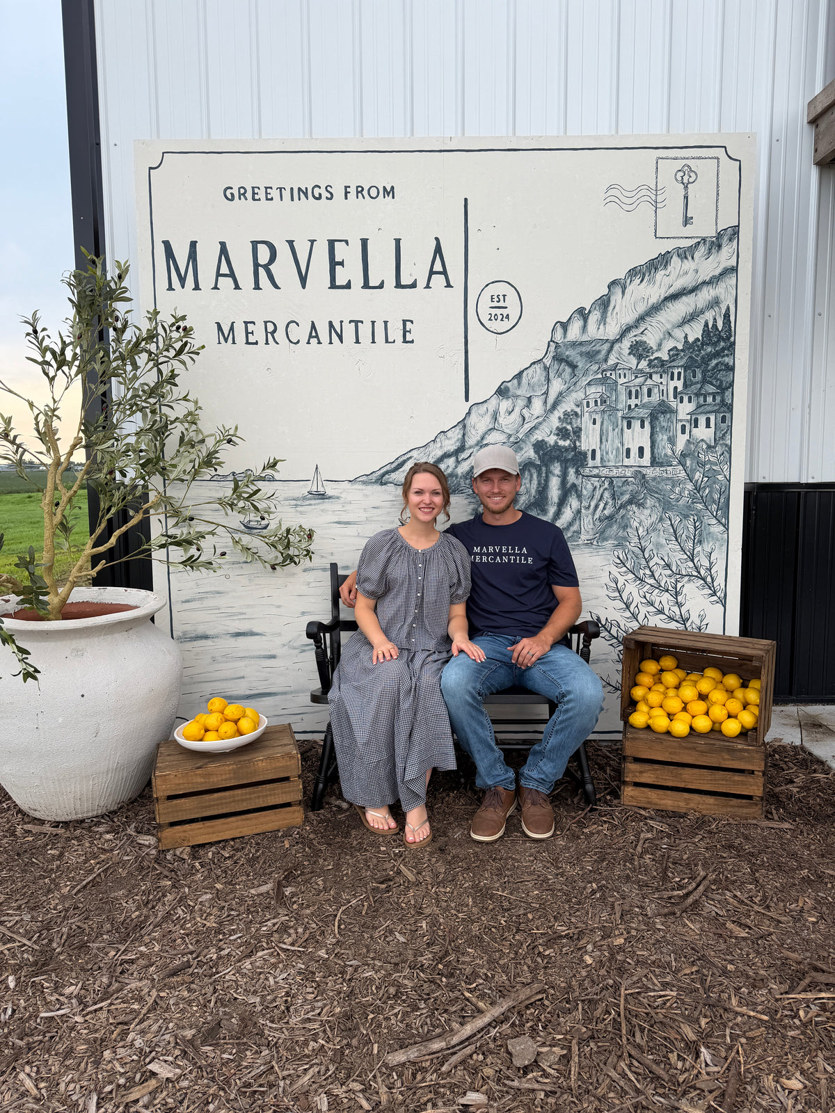 A couple sits on a bench in front of a Greetings from Marvella Mercantile postcard backdrop with crates of fresh lemons