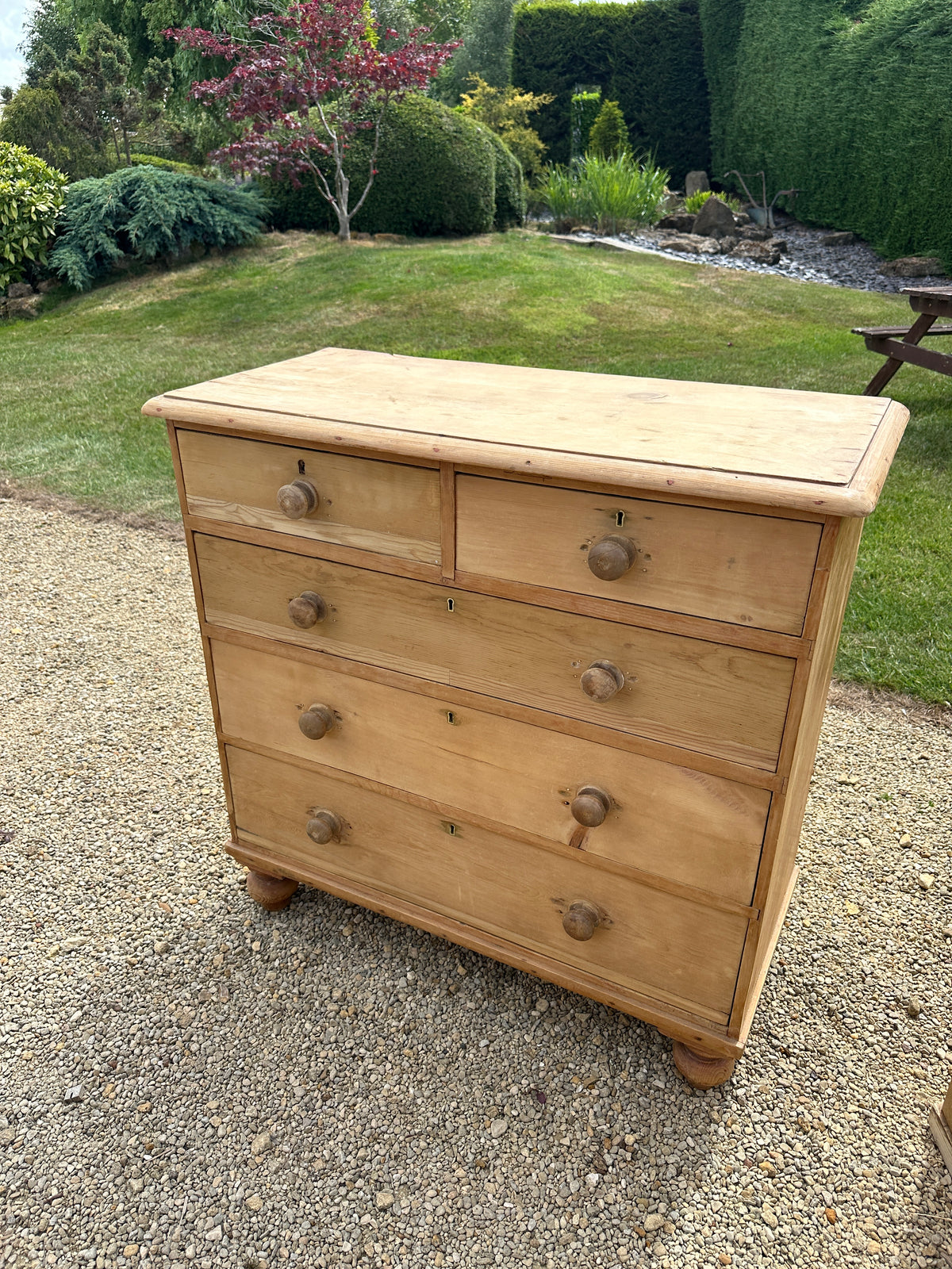 An antique English pine chest of drawers with turned wooden knobs on a gravel garden path