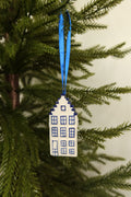 Delft blue and white ceramic Dutch canal house Christmas ornament with a stepped gable hanging from a blue ribbon on an evergreen tree