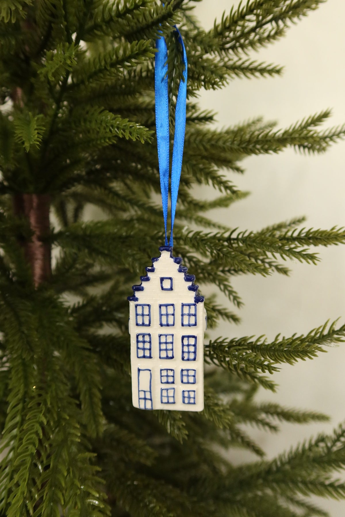 Delft blue and white ceramic Dutch canal house Christmas ornament with a stepped gable hanging from a blue ribbon on an evergreen tree