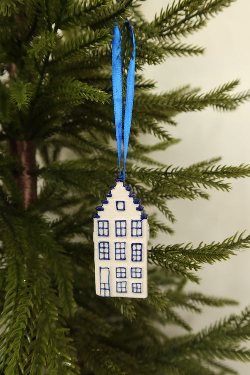 Delft blue and white ceramic Dutch canal house Christmas ornament with a stepped gable hanging from a blue ribbon on an evergreen tree