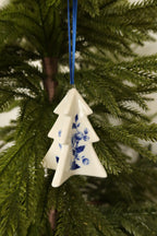 White ceramic Christmas tree ornament with a hand-painted Delft blue floral design hanging from a blue ribbon on an evergreen tree