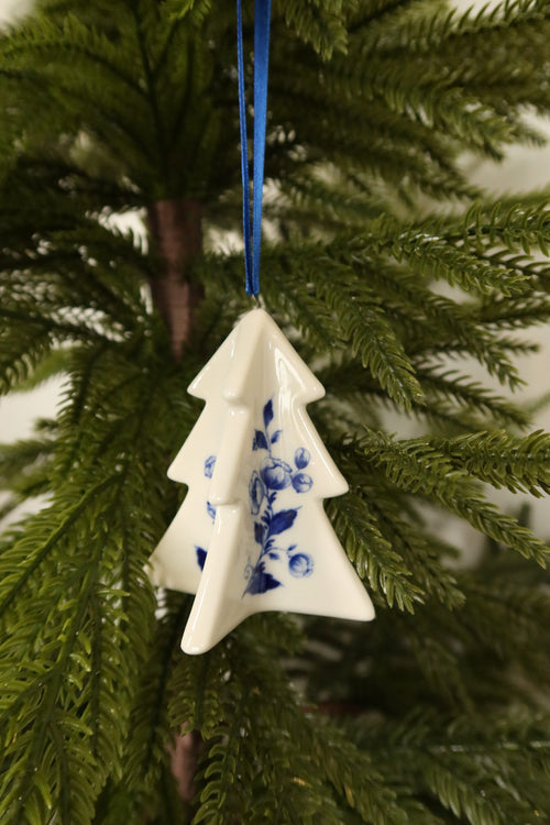 White ceramic Christmas tree ornament with a hand-painted Delft blue floral design hanging from a blue ribbon on an evergreen tree
