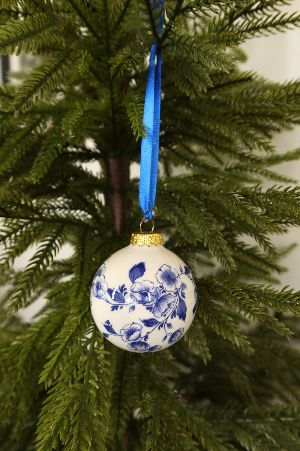 Round white ceramic Christmas bauble with a hand-painted Delft blue floral band, gold cap and blue ribbon on an evergreen tree