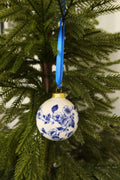 Round white ceramic Christmas bauble with a hand-painted Delft blue floral band, gold cap and blue ribbon on an evergreen tree