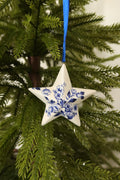 White ceramic star Christmas ornament with a hand-painted Delft blue floral design hanging from a blue ribbon on an evergreen tree