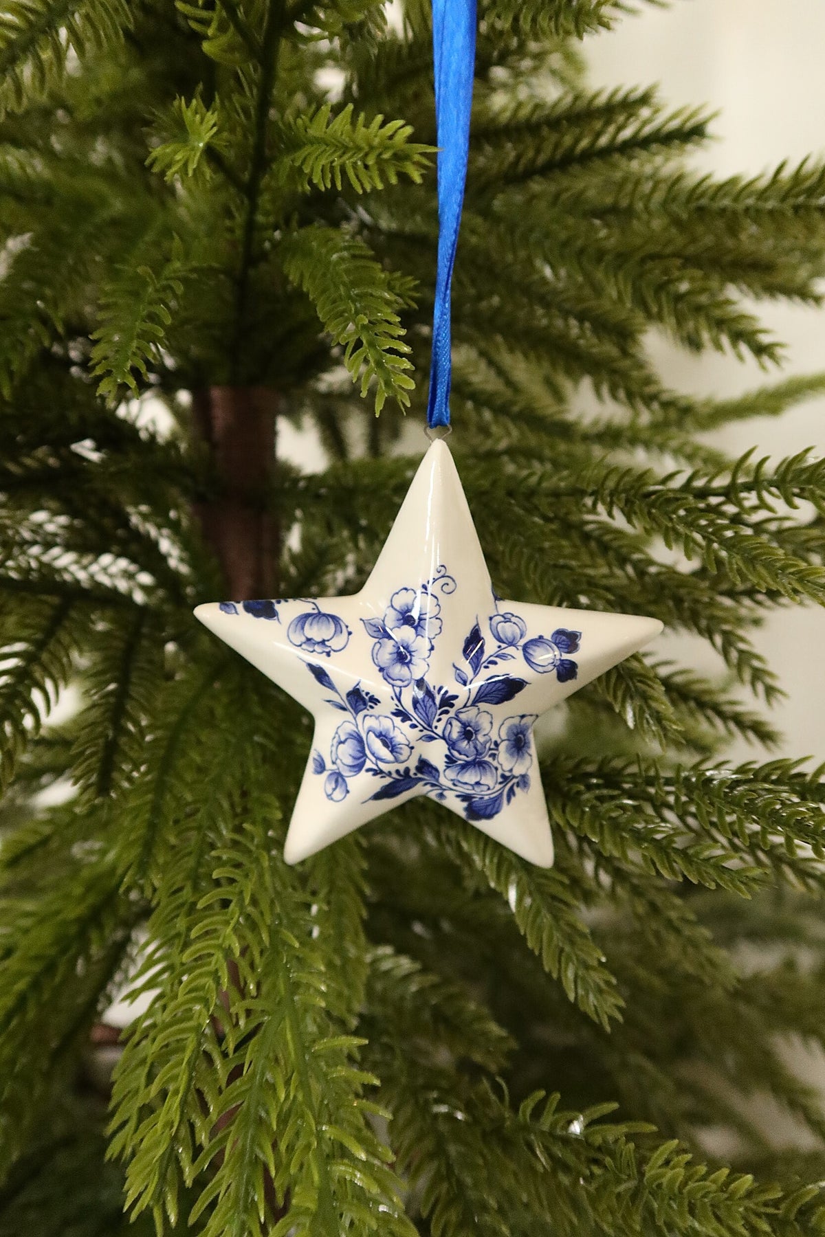 White ceramic star Christmas ornament with a hand-painted Delft blue floral design hanging from a blue ribbon on an evergreen tree