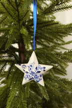 Delft blue floral star ornament on a blue ribbon among green pine branches