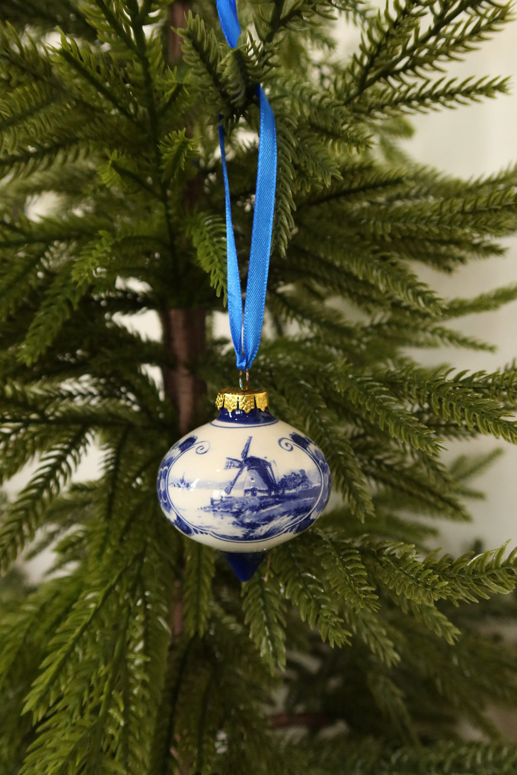 Delft blue and white ceramic drop-shaped Christmas ornament with a hand-painted windmill scene hanging from a blue ribbon on an evergreen tree