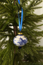 Delft blue and white ceramic drop-shaped Christmas ornament with a hand-painted windmill scene hanging from a blue ribbon on an evergreen tree