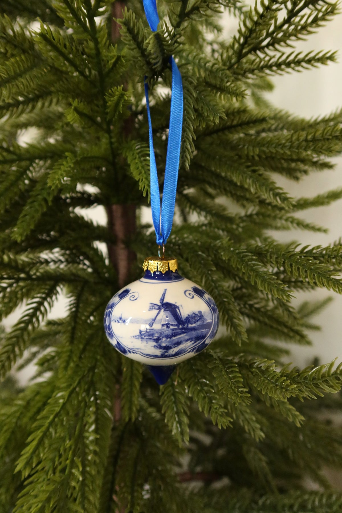 Delft blue and white ceramic drop-shaped Christmas ornament with a hand-painted windmill scene hanging from a blue ribbon on an evergreen tree