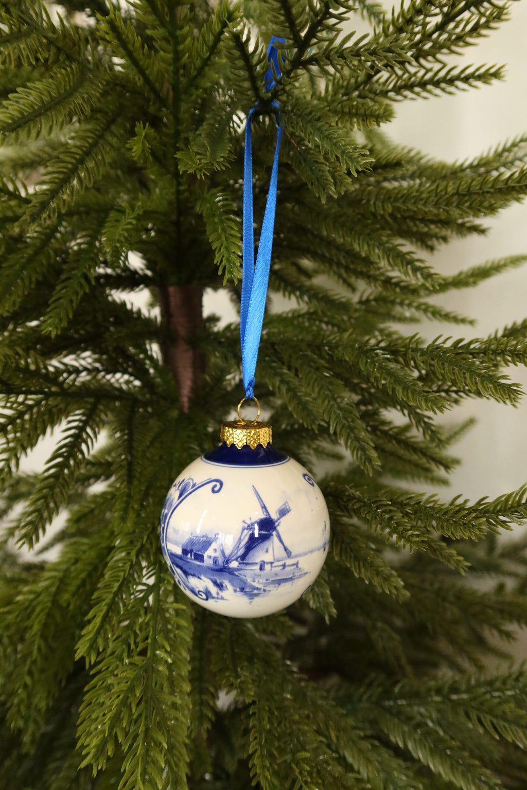 Round Delft blue and white ceramic Christmas bauble with a hand-painted windmill landscape, gold cap and blue ribbon on an evergreen tree