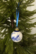 Round Delft blue and white ceramic Christmas bauble with a hand-painted windmill landscape, gold cap and blue ribbon on an evergreen tree