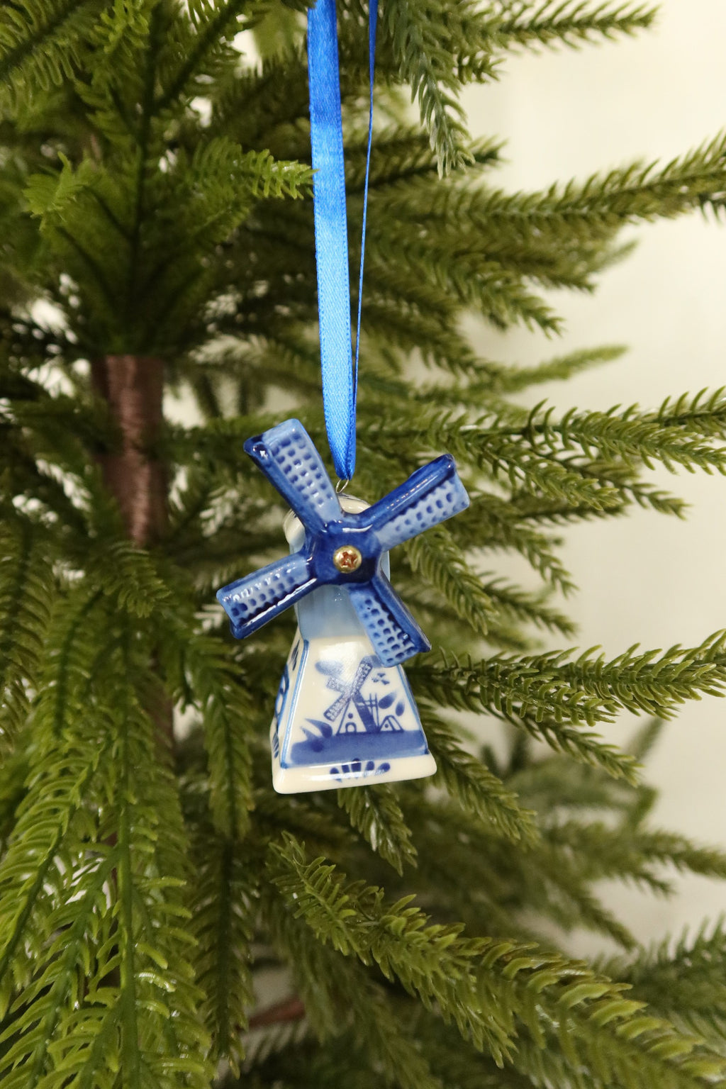 Delft blue and white ceramic windmill Christmas ornament with blue sails hanging from a blue ribbon on an evergreen tree