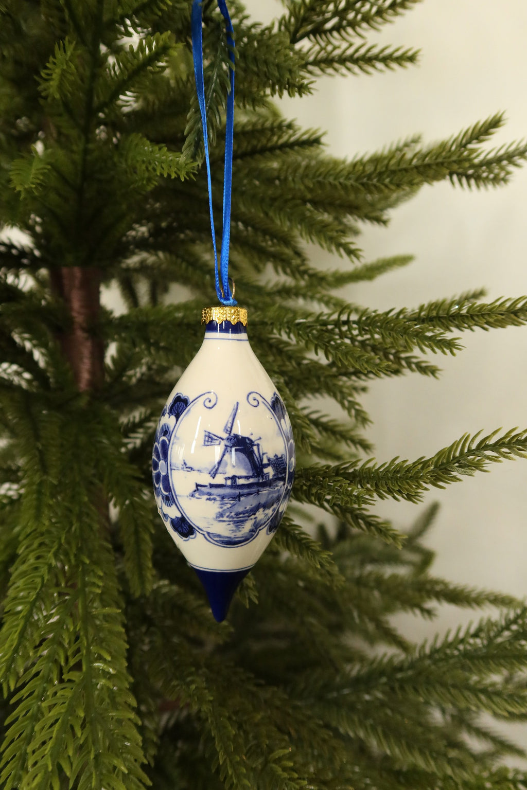 Delft blue windmill teardrop ornament with gold cap and blue tip among green pine branches
