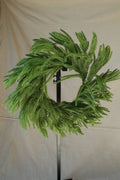 Front view of a 24 inch faux Norfolk pine wreath with long, sweeping green branches, displayed on a black stand