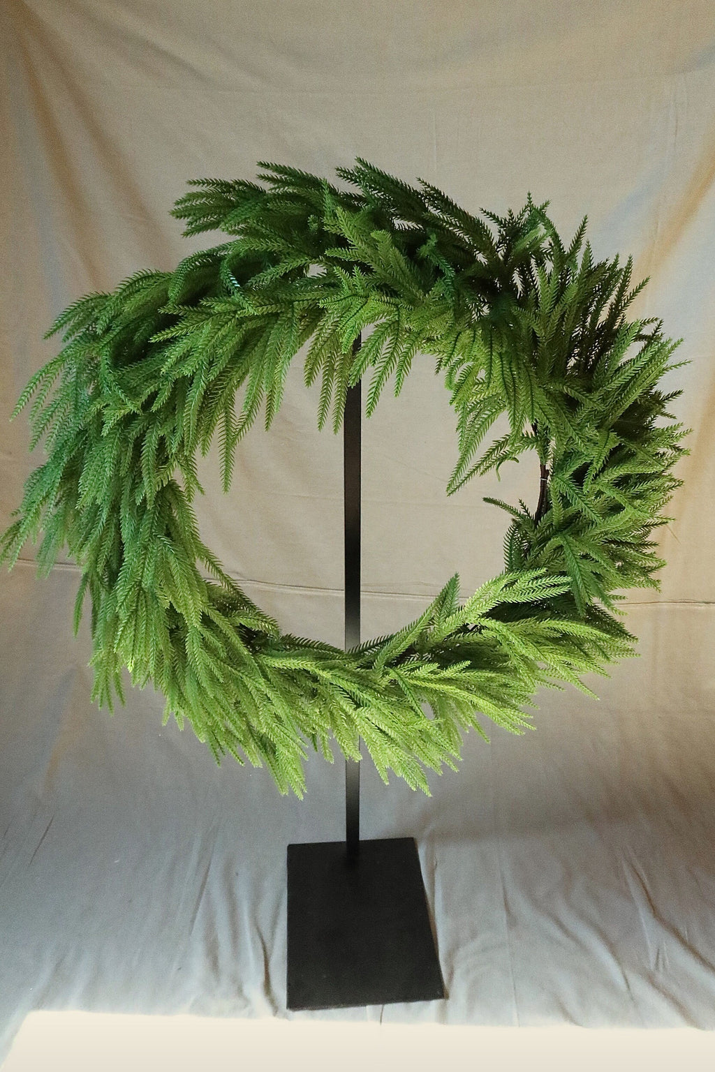 Front view of a 36 inch faux Norfolk pine wreath with long, sweeping green branches, displayed on a black stand