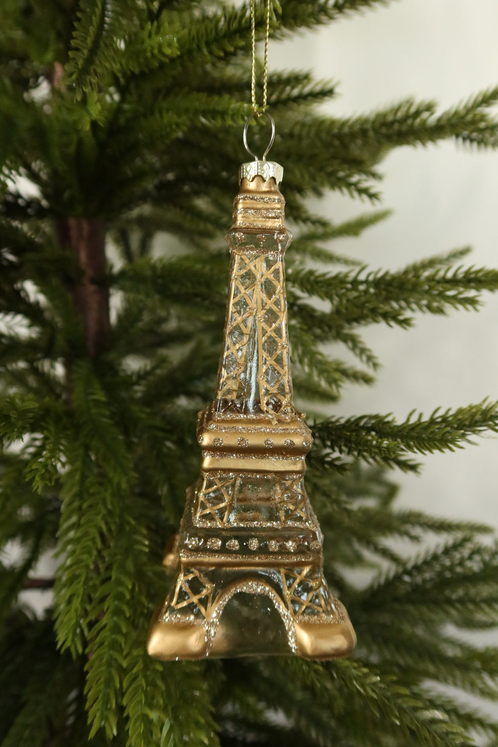Clear glass Eiffel Tower Christmas ornament with gold lattice and glitter detail hanging on an evergreen tree