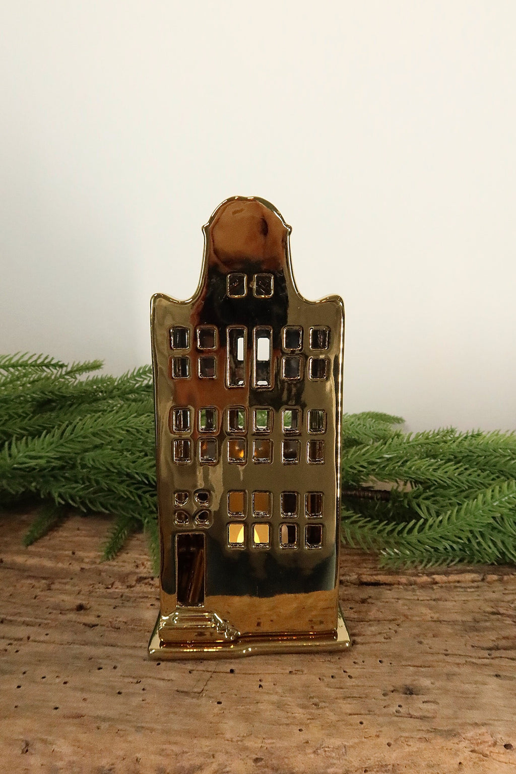 Gold ceramic canal house tealight holder with a curved bell gable, lit from within by a tealight, on a rustic wood table with greenery