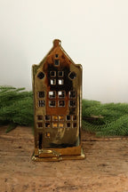 Gold ceramic canal house tealight holder with a spout gable and round attic windows, lit from within by a tealight, on a rustic wood table with greenery