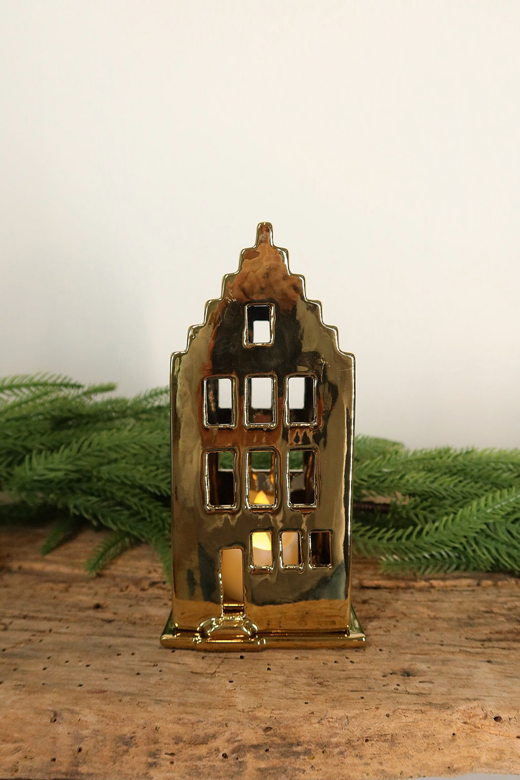 Gold ceramic canal house tealight holder with a stepped gable, lit from within by a tealight, on a rustic wood table with greenery