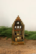 Gold ceramic canal house tealight holder with a stepped gable, lit from within by a tealight, on a rustic wood table with greenery