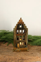 Gold ceramic canal house tealight holder with a stepped gable, lit from within by a tealight, on a rustic wood table with greenery