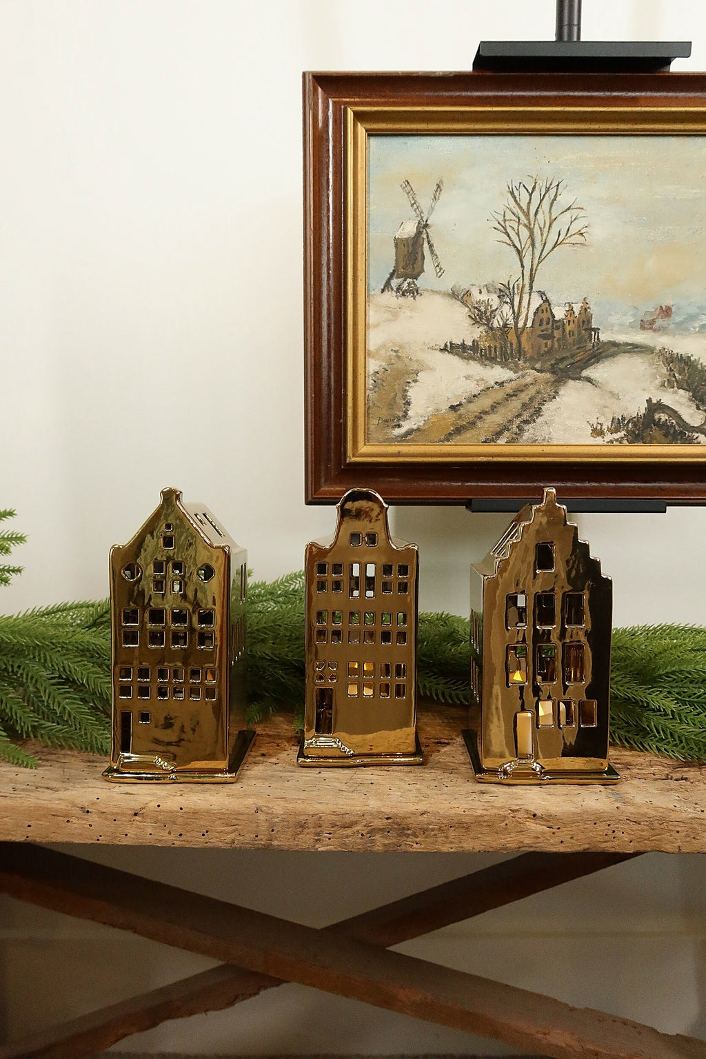Spout-gable gold canal house tealight holder on the left, shown with the bell-gable and stepped-gable houses on a rustic wood bench beneath a framed winter landscape painting