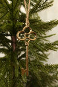 Polished gold metal skeleton key Christmas ornament with a clover-shaped bow hanging by jute twine on an evergreen tree