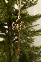 Polished gold metal skeleton key Christmas ornament with a clover-shaped bow hanging by jute twine on an evergreen tree