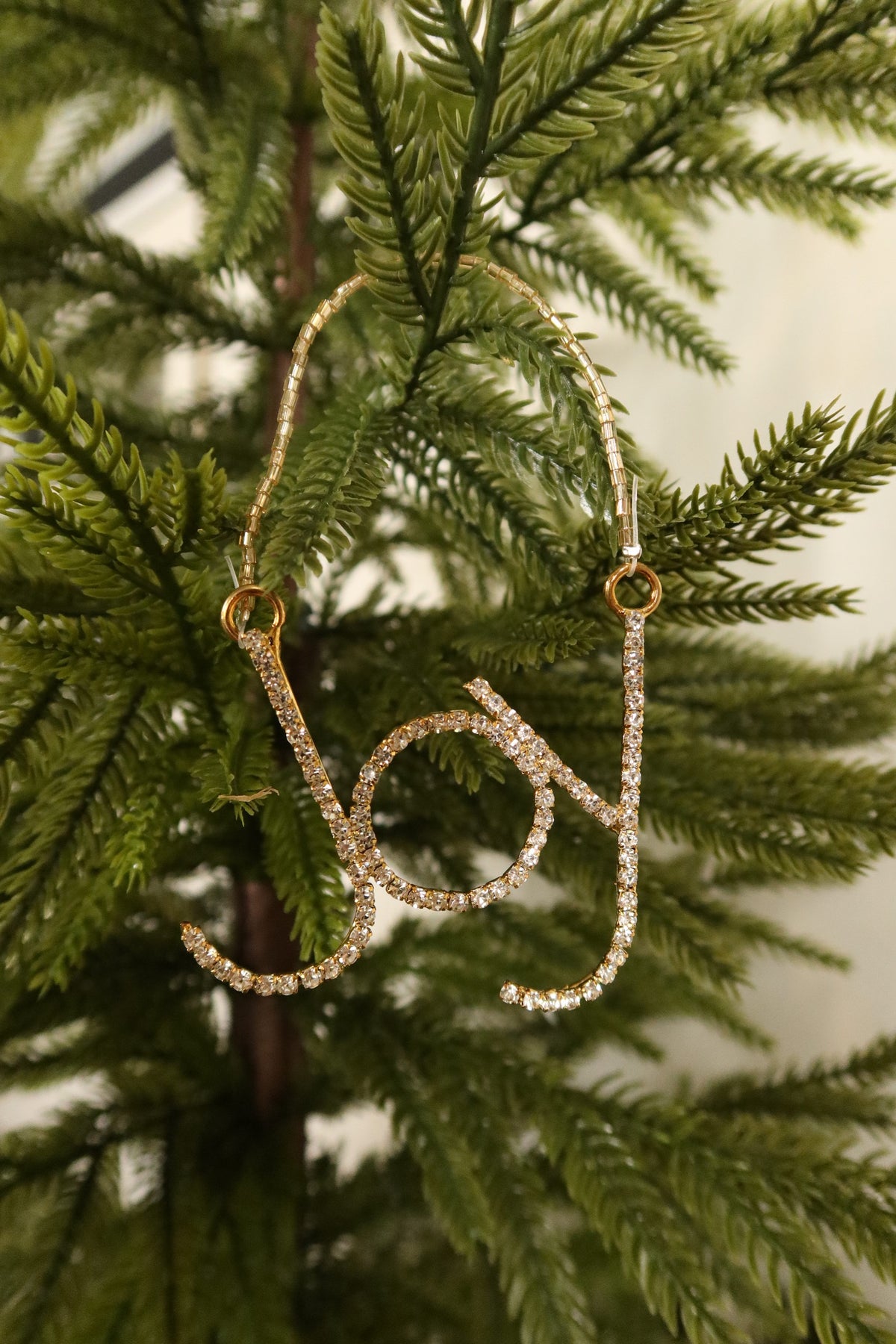 Gold rhinestone Joy script Christmas ornament hanging from a gold beaded strand on an evergreen tree