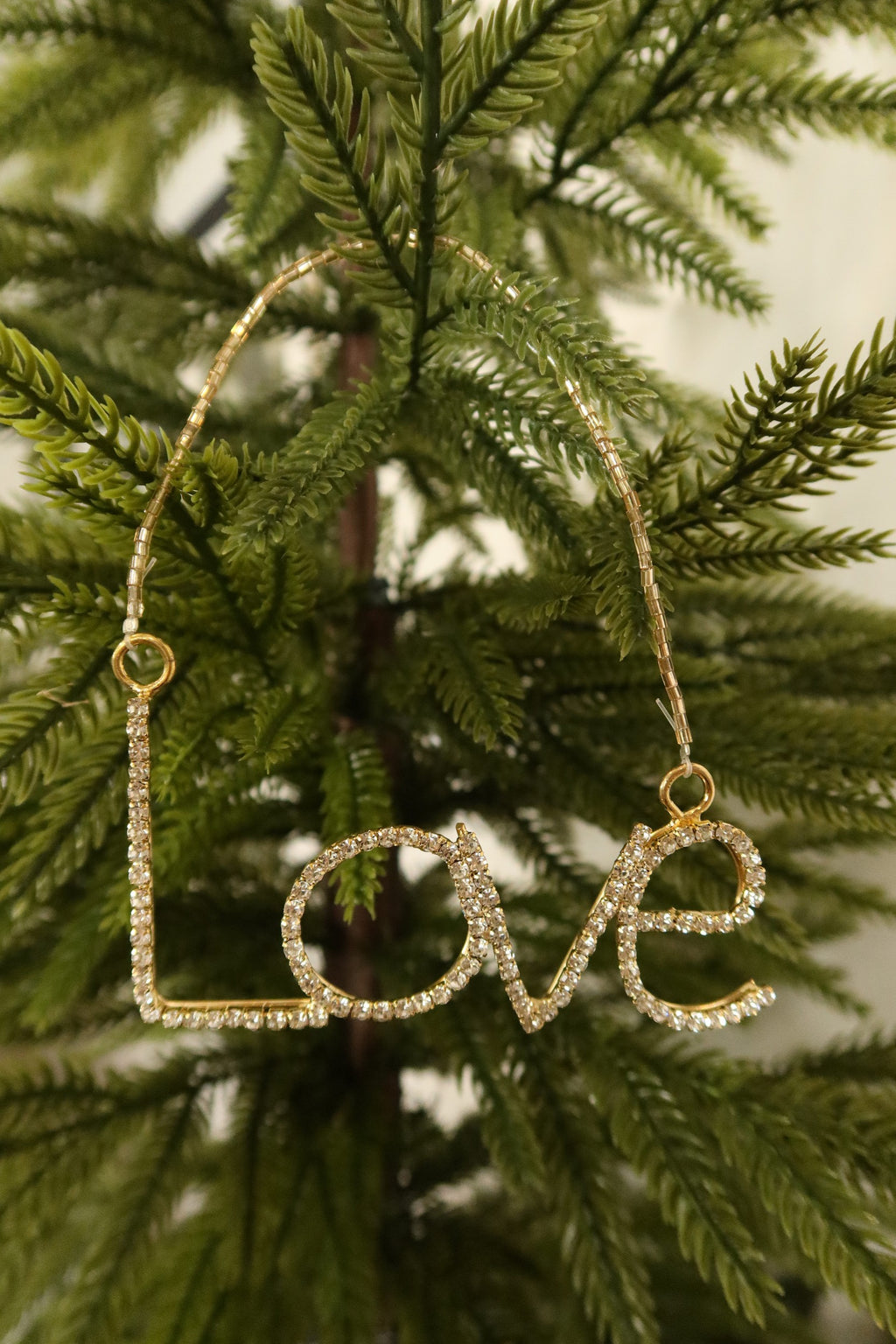 Closer view of the clear rhinestone Love ornament in a gold setting against green pine