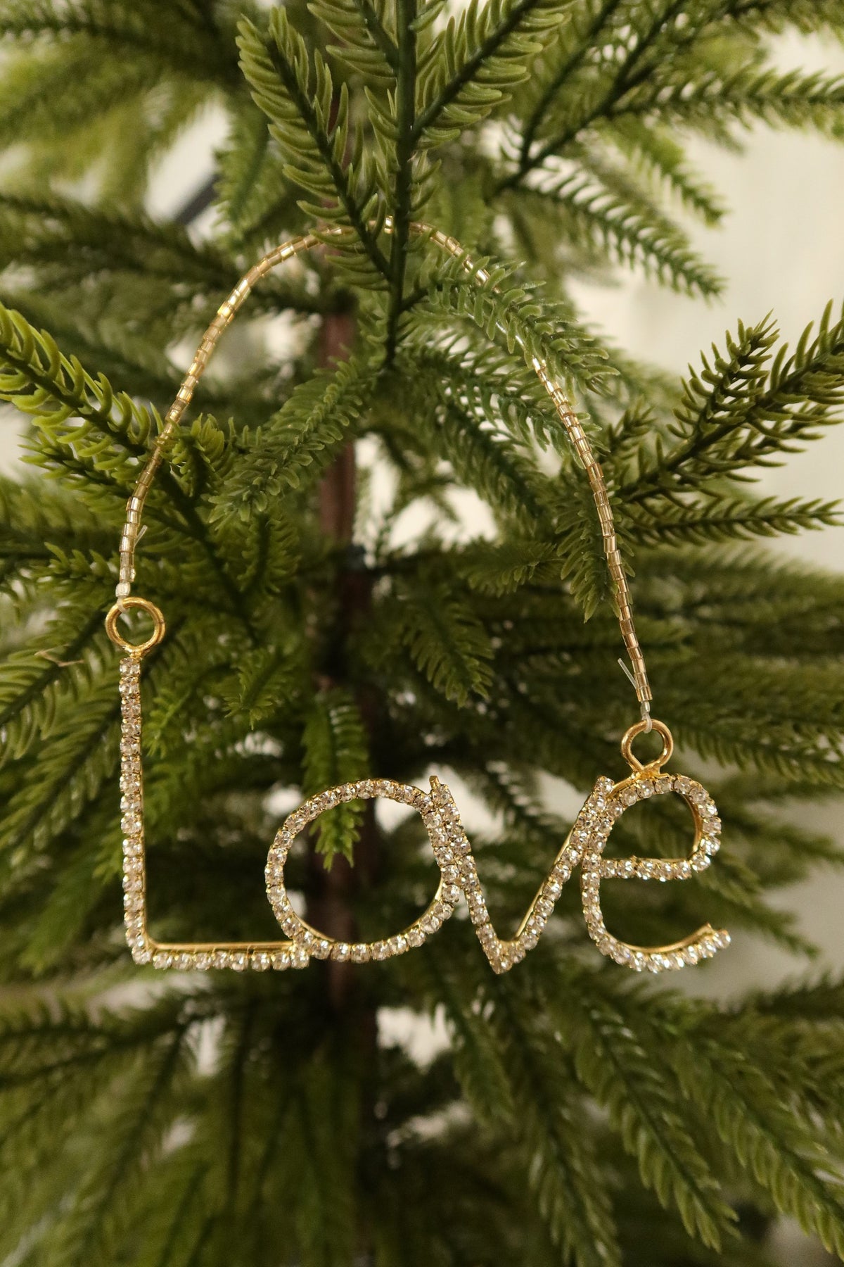 Closer view of the clear rhinestone Love ornament in a gold setting against green pine