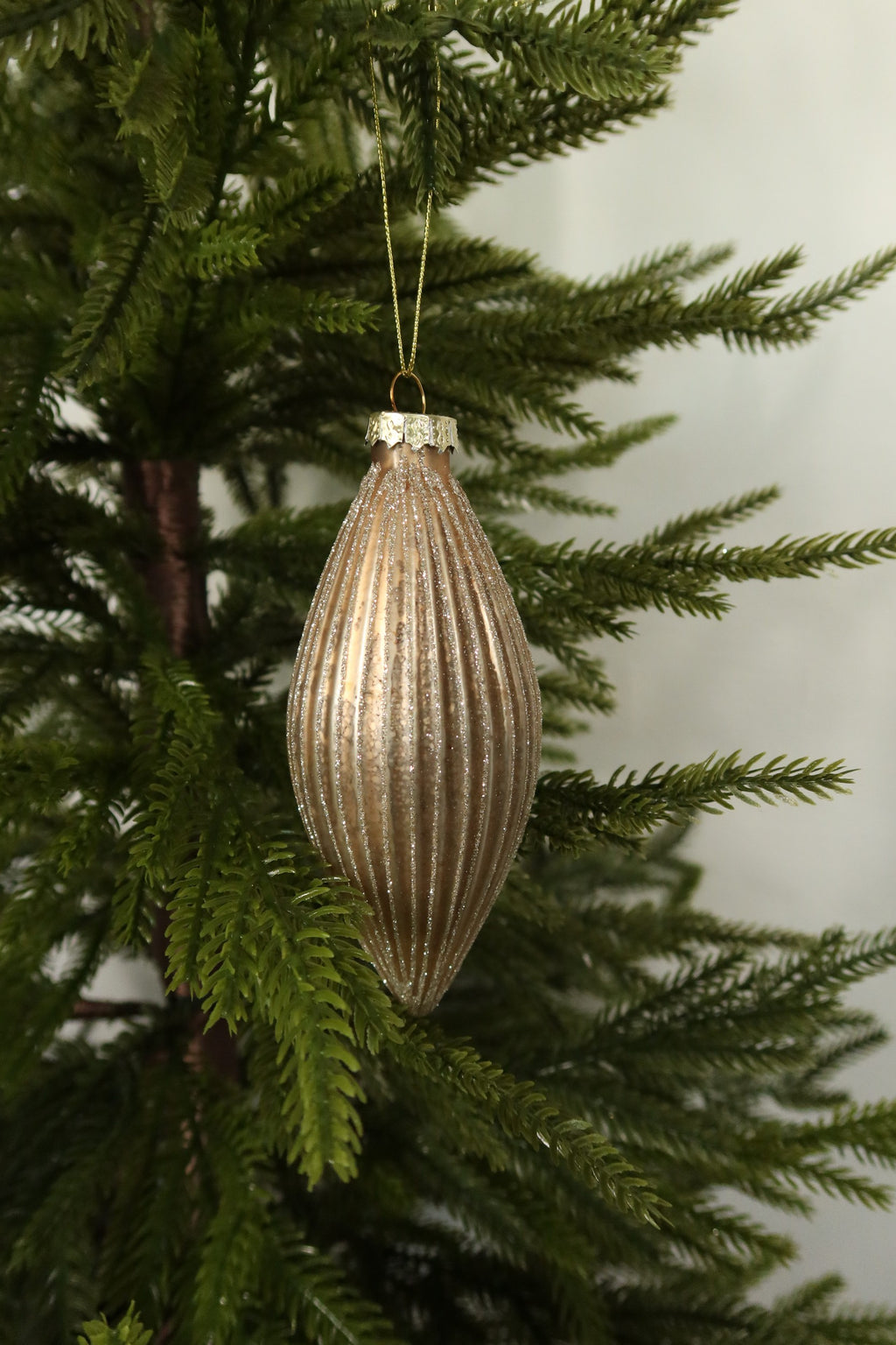 Matte gold fluted glass finial ornament with glitter-lined ribs on a gold cord among green pine branches