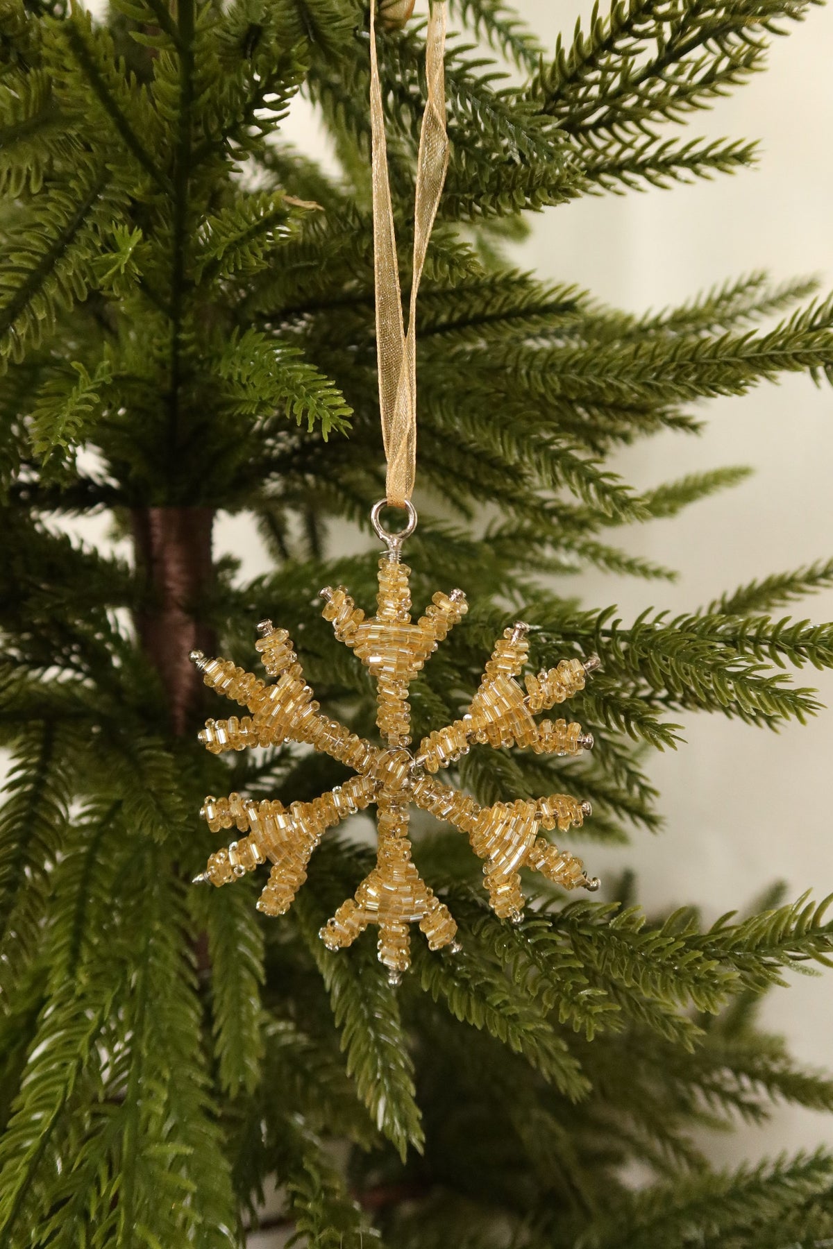 Pale gold glass bead snowflake Christmas ornament hanging from a gold ribbon on an evergreen tree