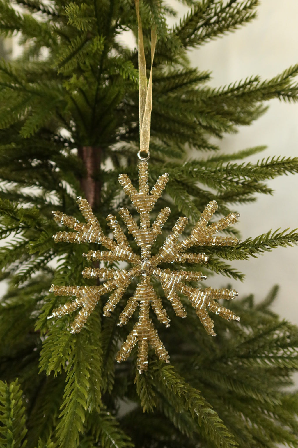 Pale gold glass bead snowflake Christmas ornament hanging from a gold ribbon on an evergreen tree