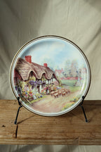 Vintage Royal Vale bone china plate with an English thatched cottage garden scene and gold rim, displayed on a black plate stand
