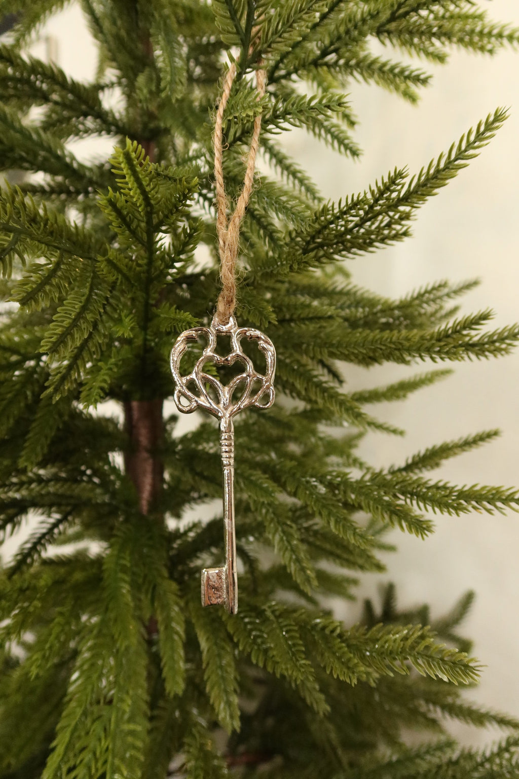 Silver metal skeleton key Christmas ornament with an ornate scrolled heart bow hanging by jute twine on an evergreen tree