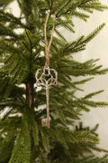 Silver metal skeleton key Christmas ornament with an ornate scrolled heart bow hanging by jute twine on an evergreen tree