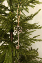 Silver metal skeleton key Christmas ornament with an ornate scrolled heart bow hanging by jute twine on an evergreen tree