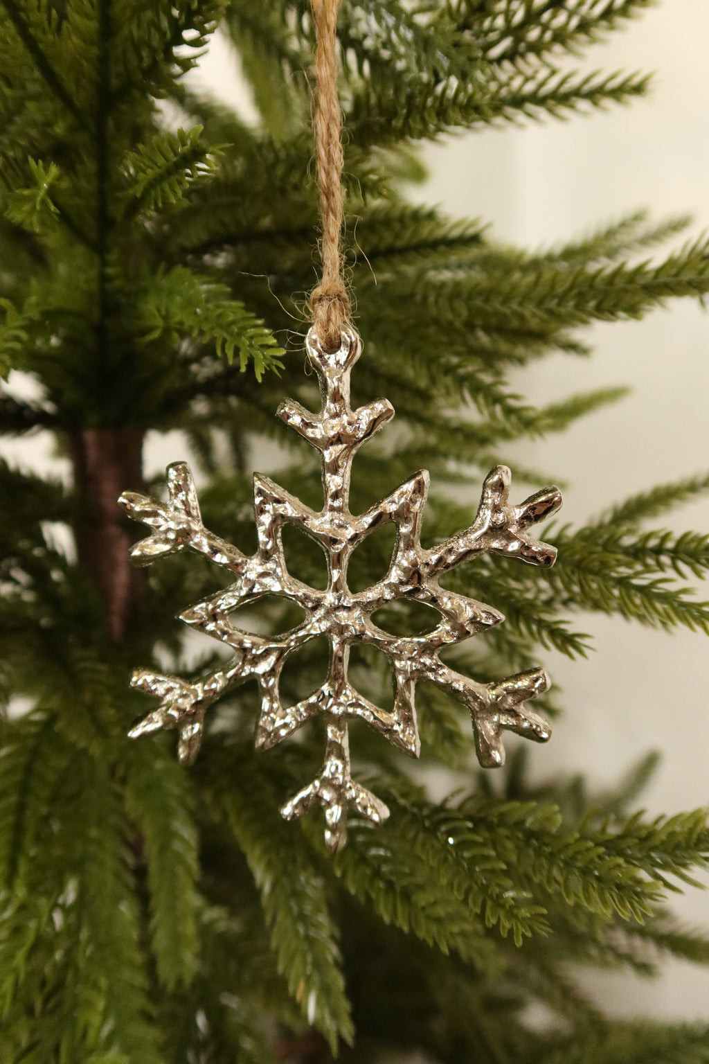 Polished silver metal snowflake Christmas ornament with a hammered texture hanging by jute twine on an evergreen tree