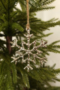 Polished silver metal snowflake Christmas ornament with a hammered texture hanging by jute twine on an evergreen tree