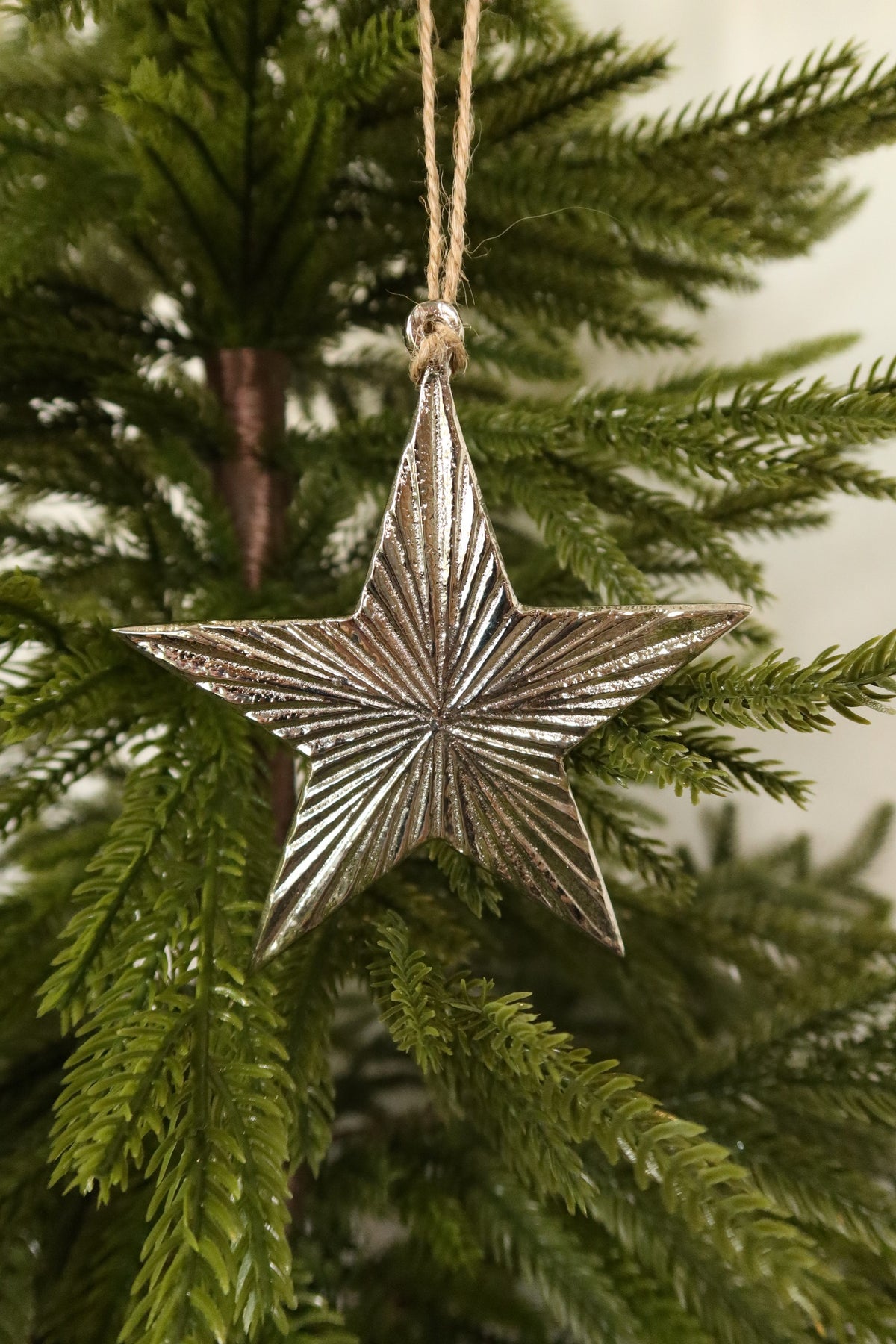 Silver metal star Christmas ornament with ridged radiating texture hanging by jute twine on an evergreen tree