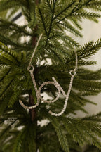 Silver rhinestone Joy script Christmas ornament hanging from a silver beaded strand on an evergreen tree