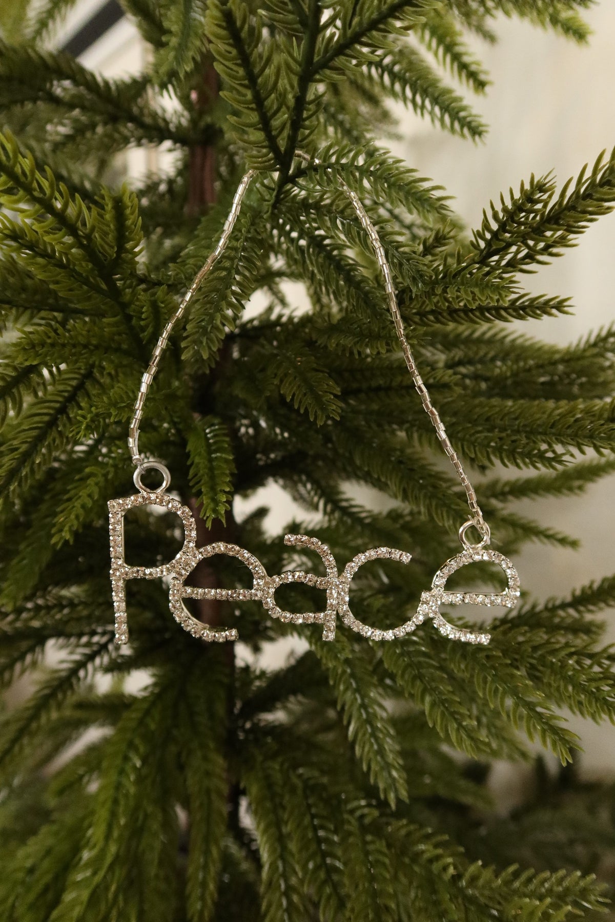 Silver rhinestone Peace word Christmas ornament hanging from a silver beaded strand on an evergreen tree