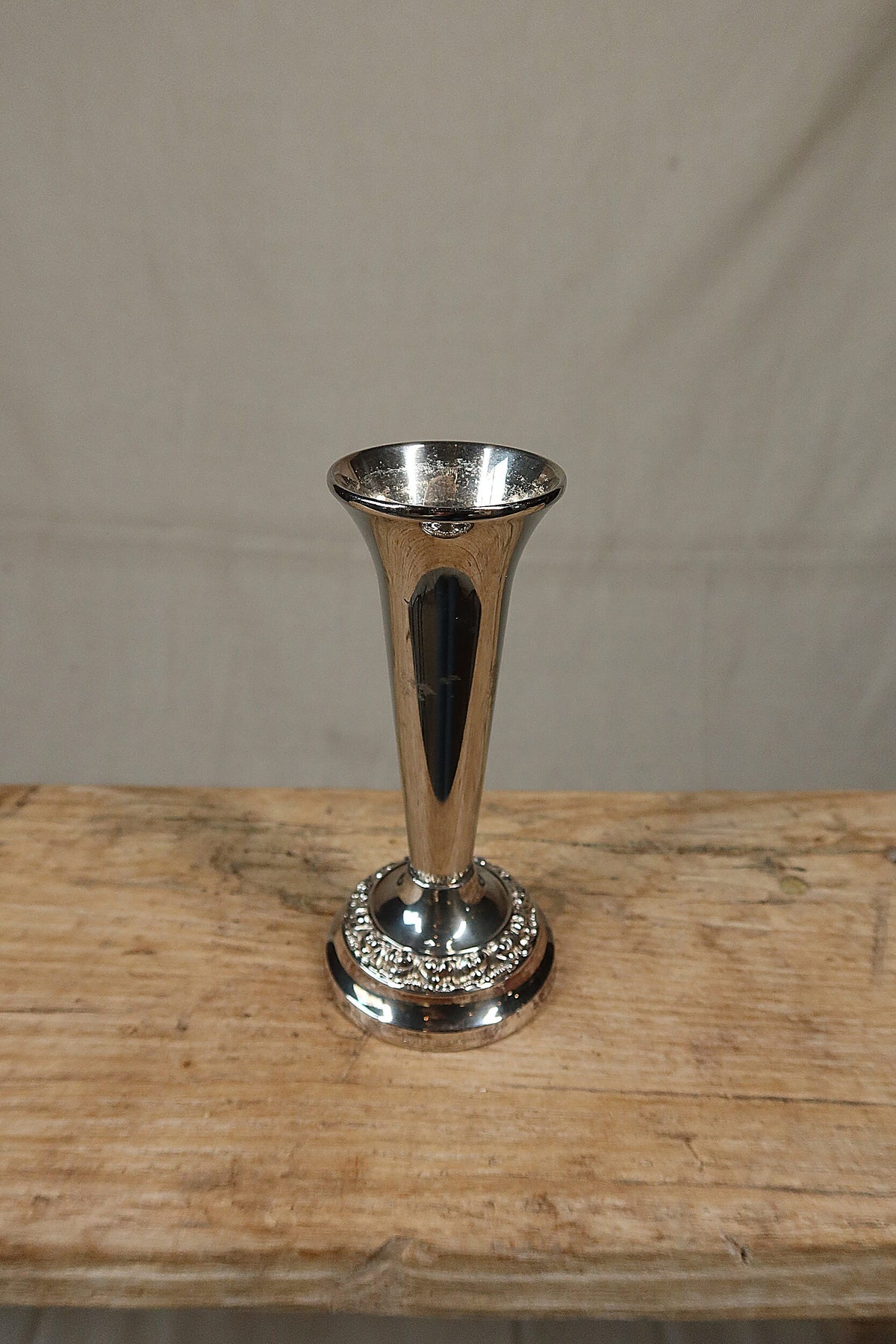 Vintage Ianthe of England silver plate bud vase with a flared trumpet neck and embossed floral foot, standing on a rustic wooden bench